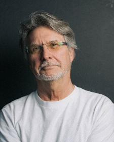 Headshot of grey-haired man in white tshirt leaning against a grey wall.